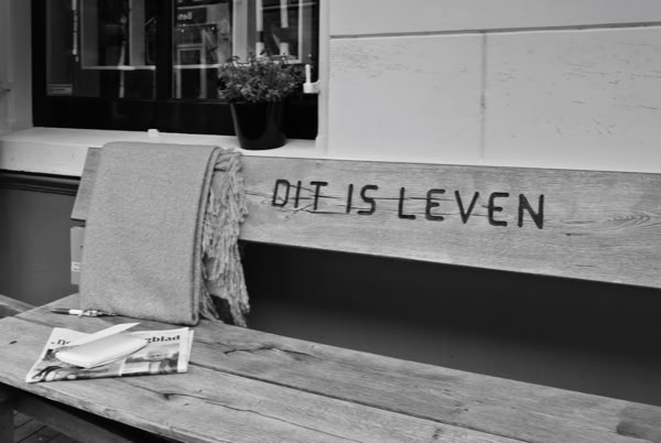 A wooden bench with a blanket draped over one side and a newspaper on the seat. The words "DIT IS LEVEN" are carved into the backrest. A small plant is visible in the background.