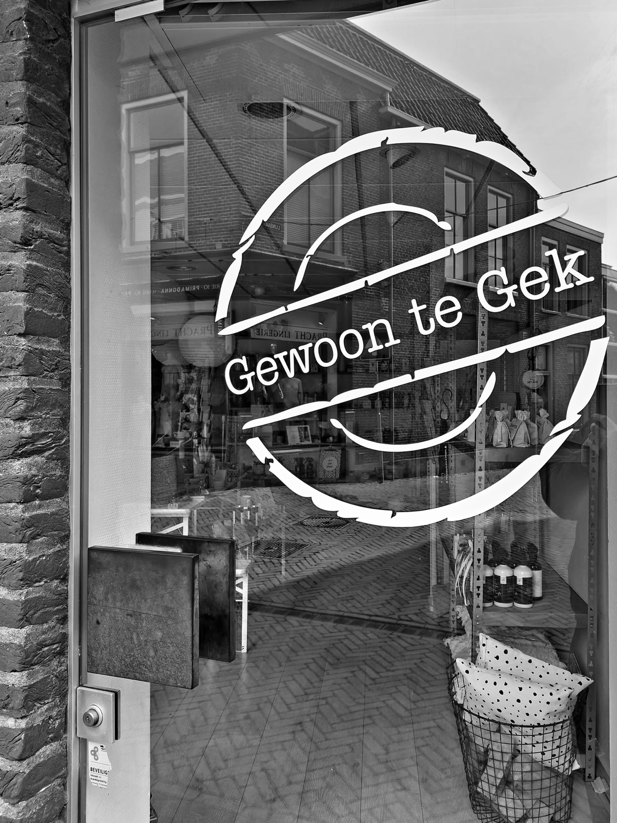 A glass door with the text "Gewoon te Gek" (Simply to crazy) in a circular design, reflecting a street scene with buildings.