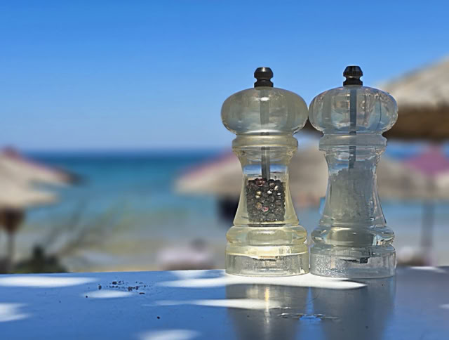 Two clear salt and pepper grinders on a table with a blurred beach and umbrellas in the background.