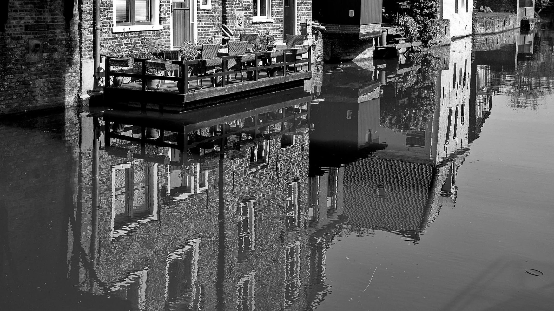 A black and white image of a brick building with a wooden deck, reflected in a body of water.