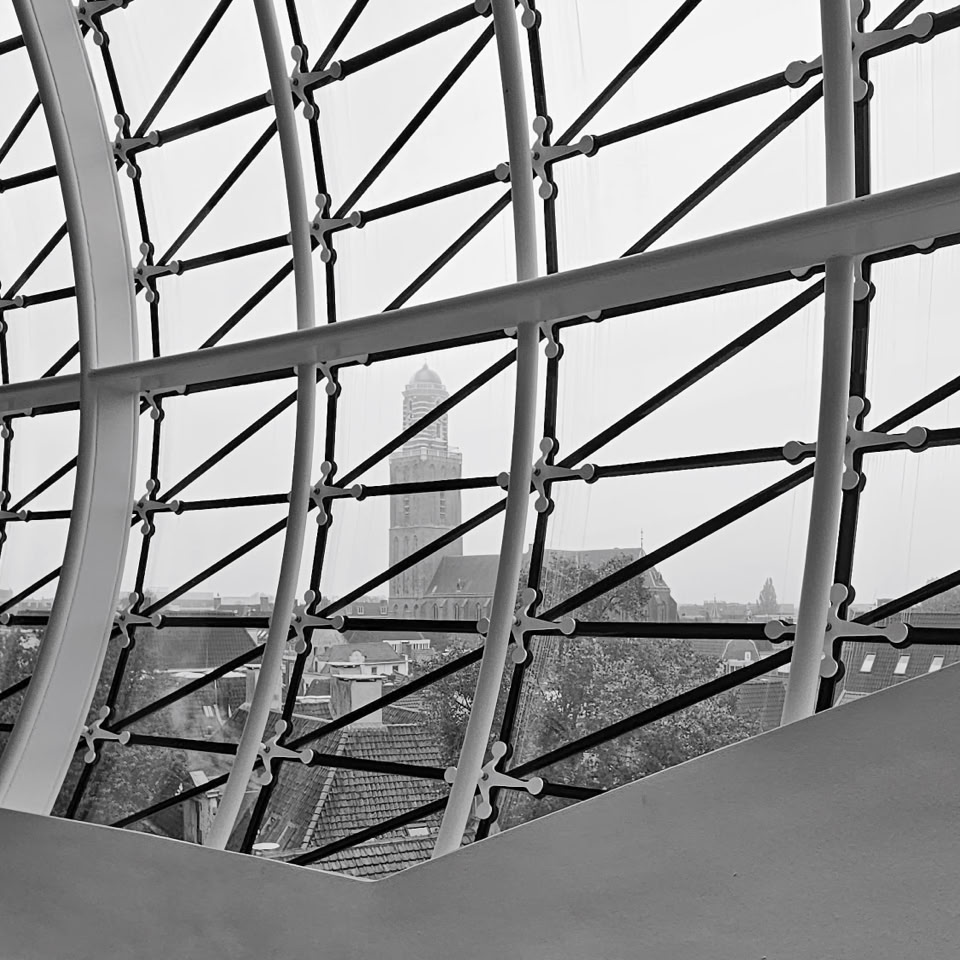Curved metal and glass structure with a view of a distant clock tower and buildings. Zwolle, Netherlands