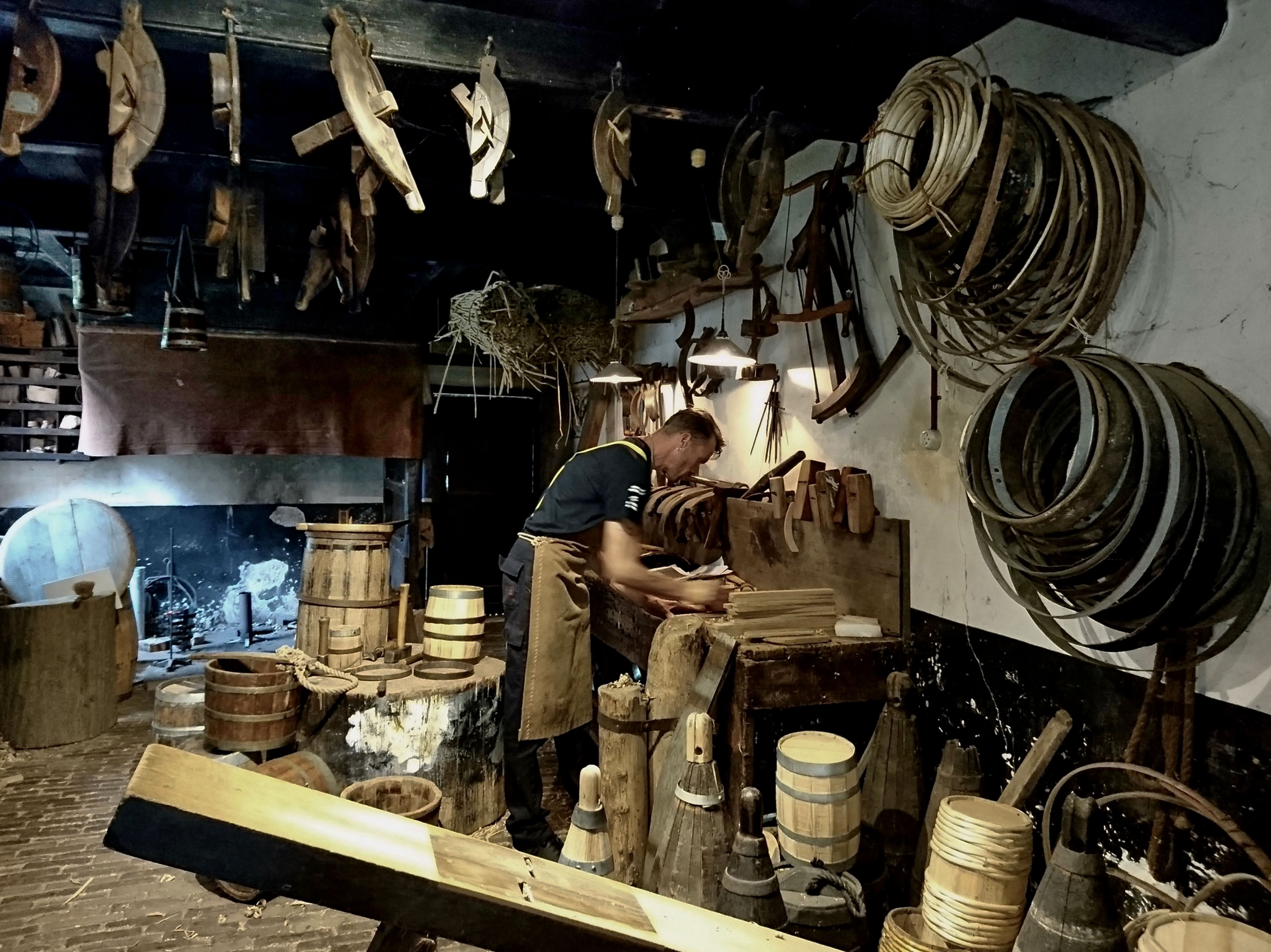 A person working in a workshop filled with wooden barrels, tools, and hanging items, with a fireplace in the background.