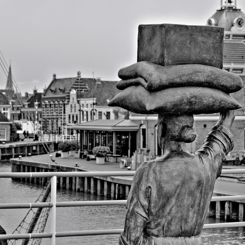 A statue of a person carrying bags on their head, overlooking a waterfront with buildings and a bridge in the background. The image is in black and white. Harlingen, Netherlands