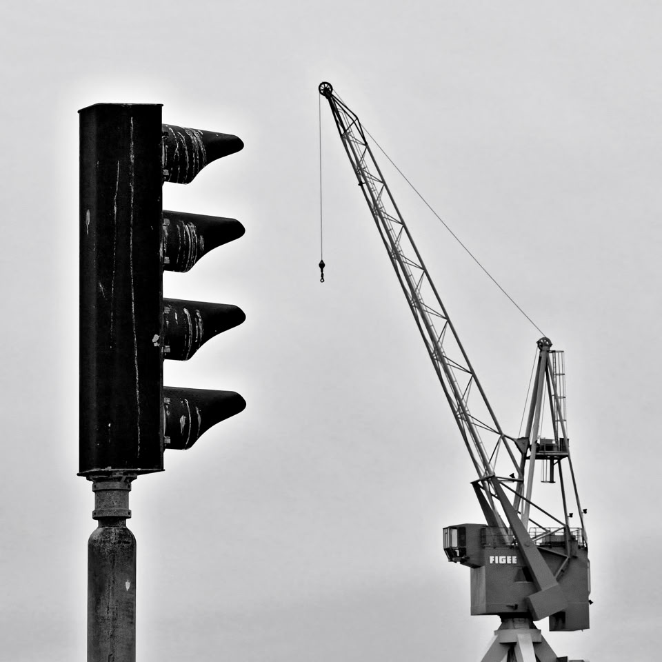 A black and white image showing a traffic light on the left and a large crane on the right against a clear sky. Harlingen, Netherlands