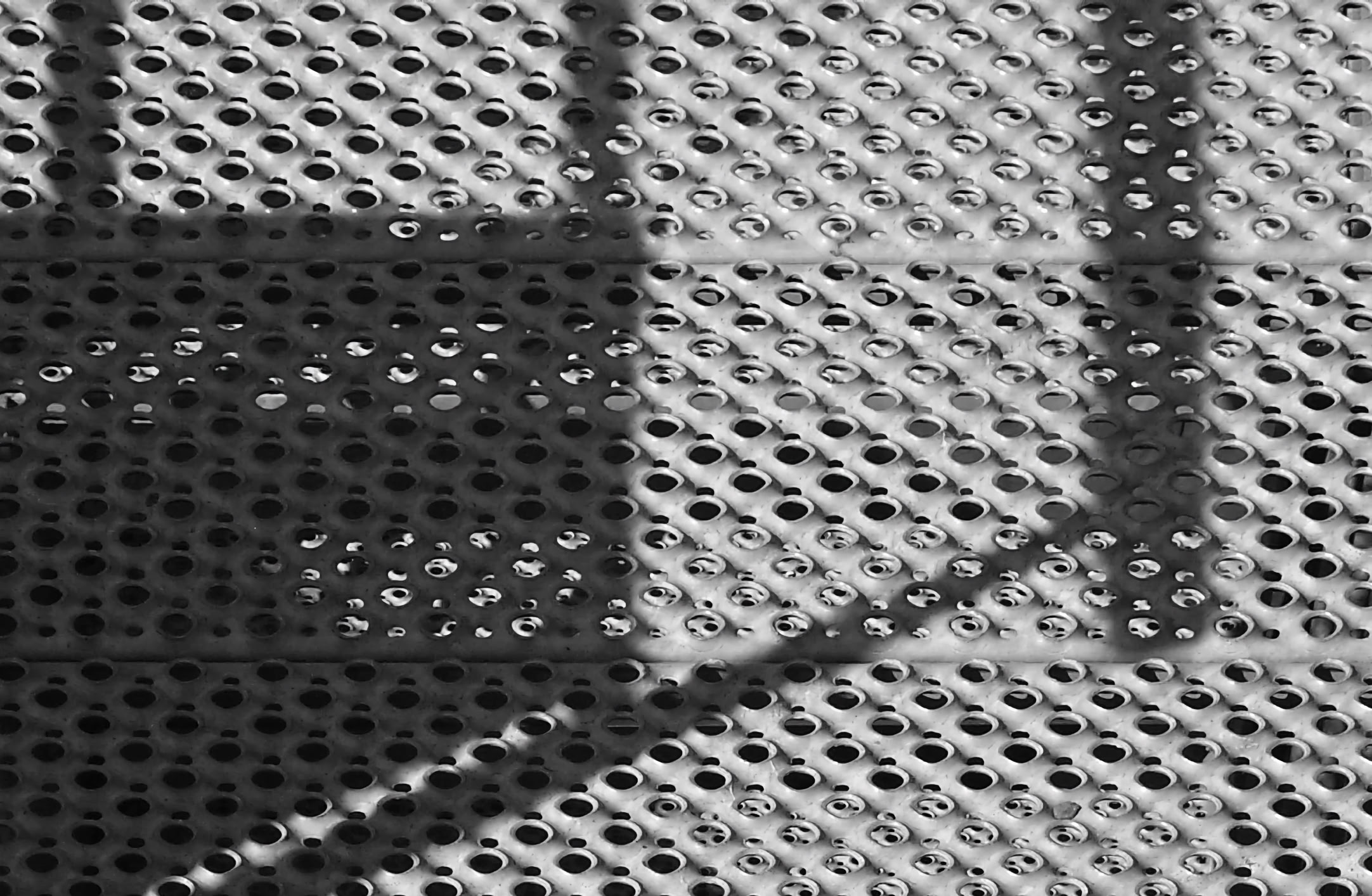 Black and white image of a perforated metal surface with shadows creating geometric patterns.
