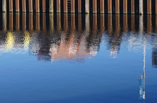 Reflection of colorful buildings and a mast in calm water.