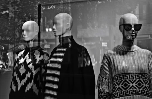 Three mannequins wearing patterned sweaters are displayed in a store window. The mannequin on the right is wearing sunglasses.